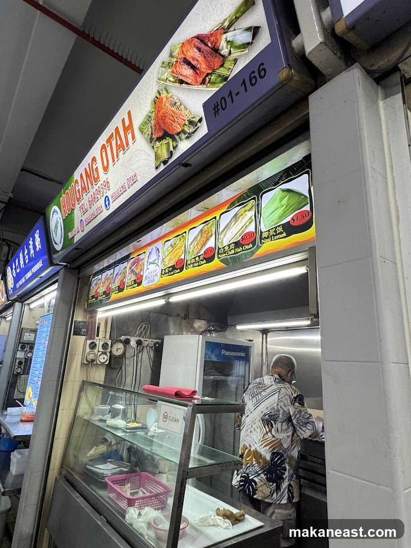 A food stall with a prominent sign displaying "HOUGANG OTAH" and images of otah. Below the main sign, a menu board shows various food items with prices. A person is visible working behind the counter.