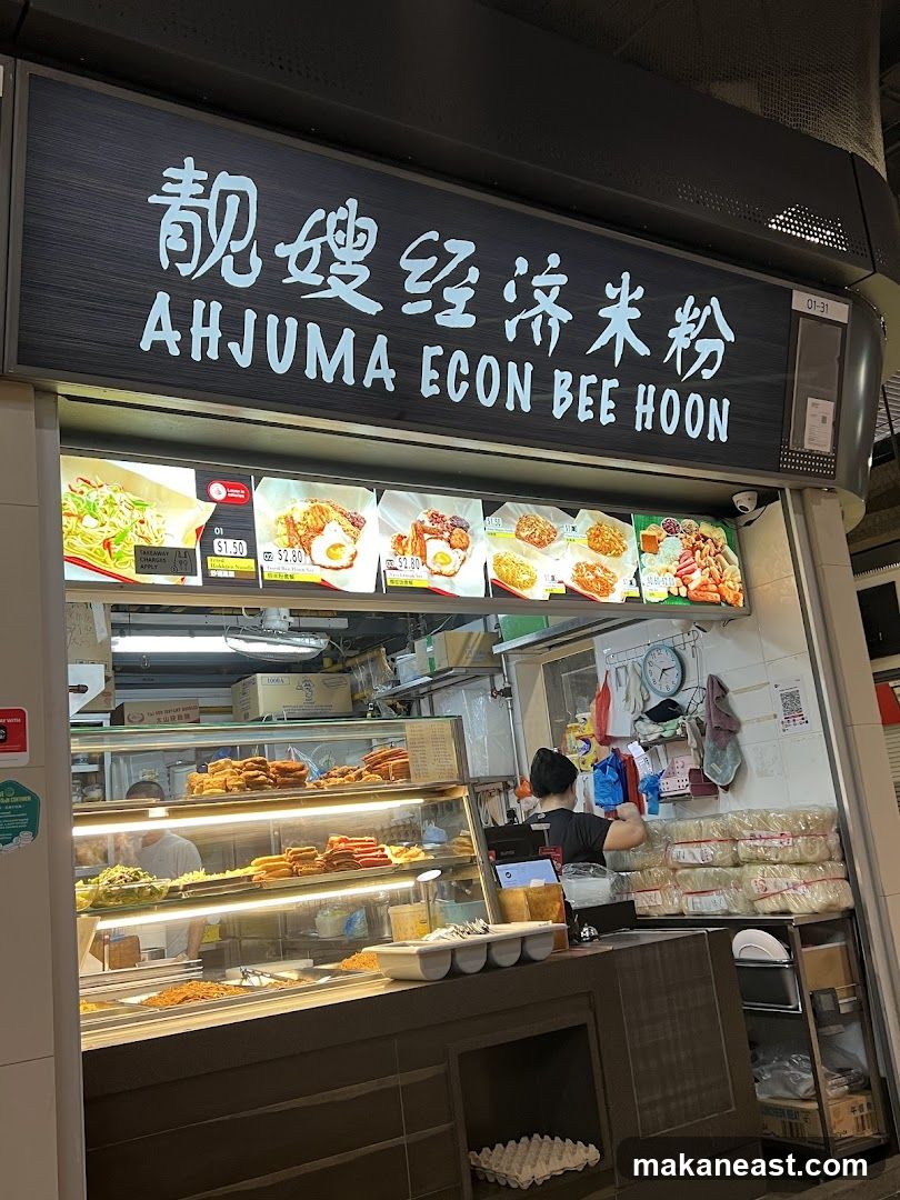 A food stall with a sign that reads "AHJUMA ECON BEE HOON" in English and Chinese. Below the sign are pictures of various dishes with prices. The stall displays food items in a refrigerated case, and a person is working behind the counter.