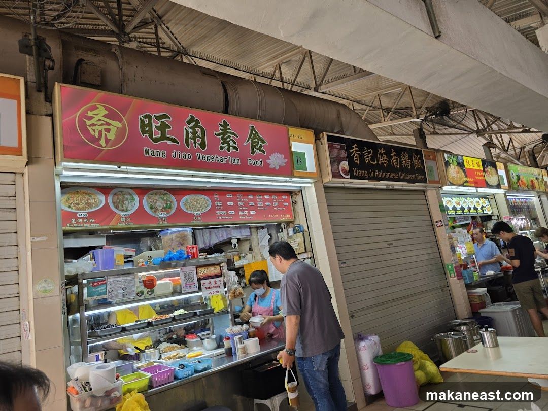 A food stall with a large red sign that reads "Wang Jiao Vegetarian Food" in Chinese and English. Below the sign are pictures of various dishes with prices. The stall displays a variety of food items in refrigerated cases. Two staff members are visible behind the counter, and customers are ordering food. Other food stalls are visible to the right.