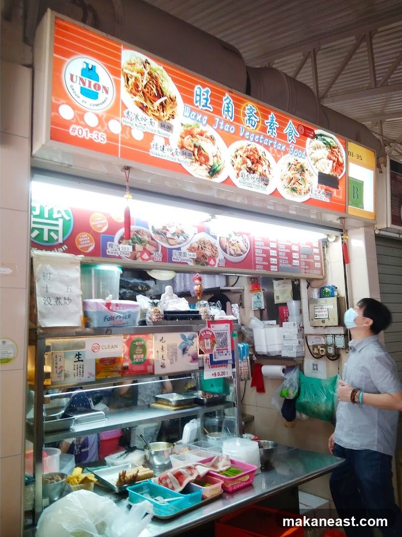 A street food stall with a large illuminated sign displaying food items and prices. The stall has a display case with various food ingredients and prepared dishes. A person wearing a mask is looking at the menu.