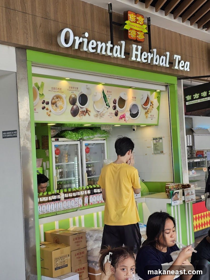 A brightly lit shop front for Oriental Herbal Tea, featuring a prominent sign with the business name. Below the sign is a colorful display showcasing various herbal drinks and desserts with accompanying images and text. The shop has refrigerators stocked with bottled drinks and a counter where staff can serve customers. Several people are visible, including staff and customers enjoying drinks. Boxes are stacked near the entrance.