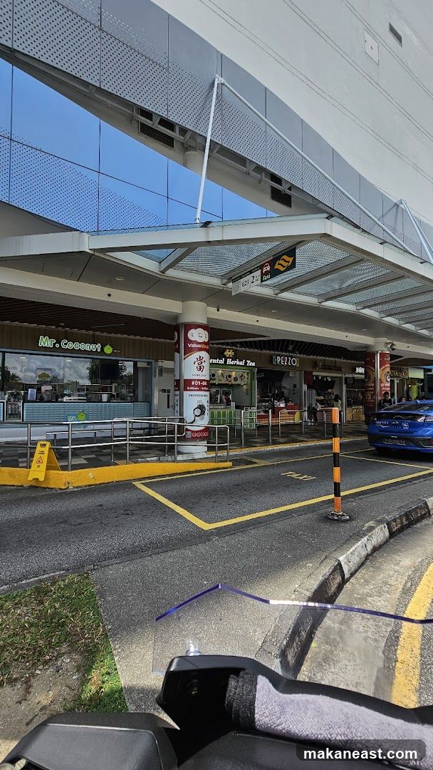 A row of shops with awnings and signage is visible from a street-level perspective. The shops include "Mr. Coconut", "ValueMalt", "Oriental Herbal Tea", and "PEZZO". A blue taxi is parked in the foreground. The building facade above the shops features reflective blue glass and a modern, perforated metal design.