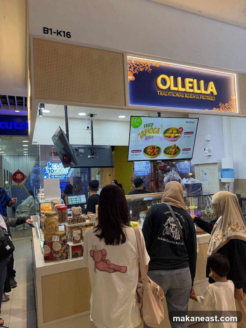 A storefront for Ollella, a shop selling traditional kueh and pastries. The shop has a prominent sign with the name OLLELLA in gold lettering. Below the sign, a digital menu displays images and prices of fried tapioca balls and other items. Customers are seen queuing and browsing the products displayed in glass cases.
