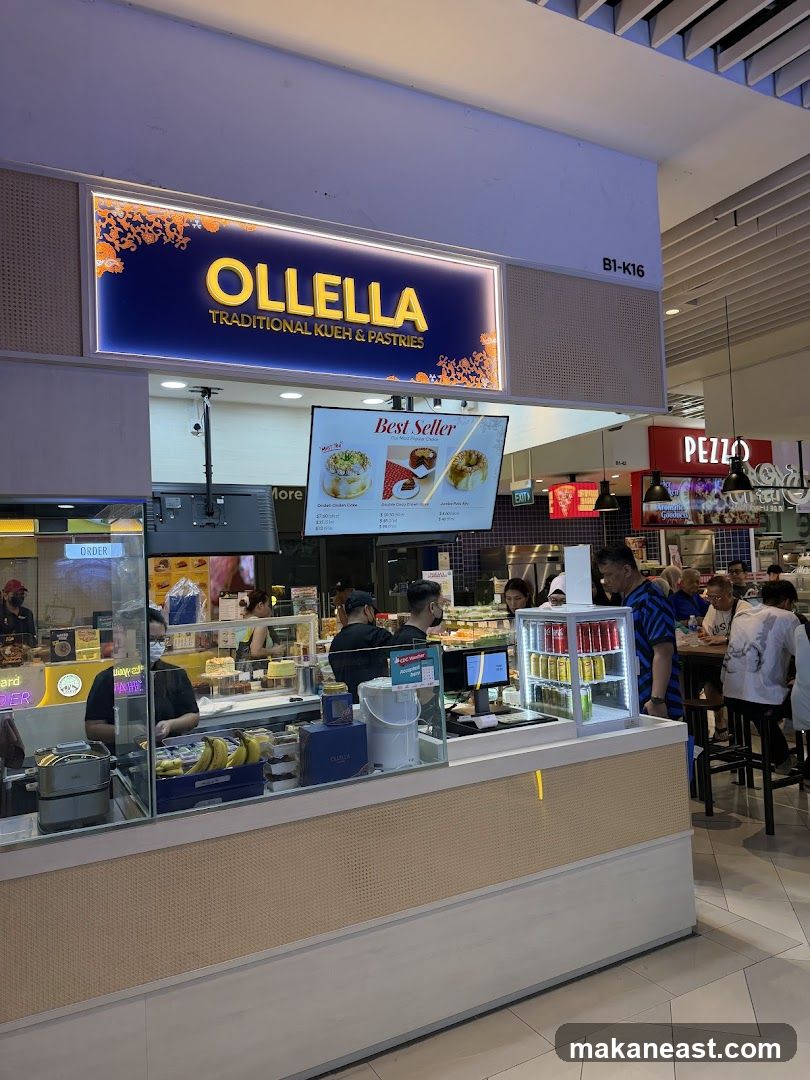 The exterior of a food stall with a prominent sign that reads "OLLELLA TRADITIONAL KUEH & PASTRIES". Below the sign is a digital menu displaying "Best Seller" items. The stall has a counter with various pastries and drinks, and staff members are serving customers. Other food establishments are visible in the background.