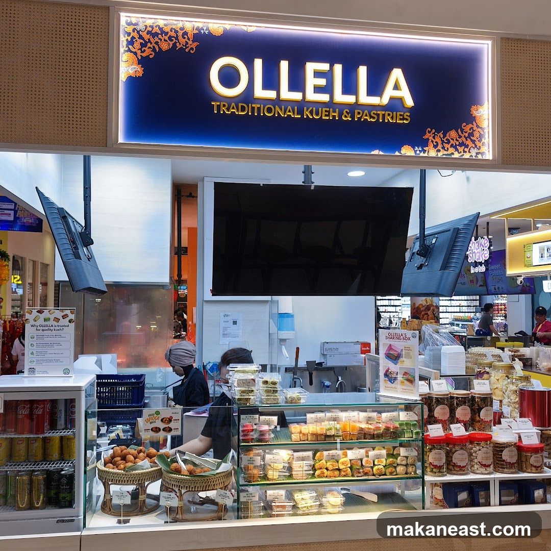 A brightly lit storefront for Ollella, a shop specializing in traditional kueh and pastries. The shop's name and slogan are prominently displayed on a large illuminated sign above the counter. The display counter features a variety of colorful pastries and snacks in individual packaging, as well as jars of nuts and cookies. A refrigerator stocked with drinks is visible to the left. Two staff members are working behind the counter.