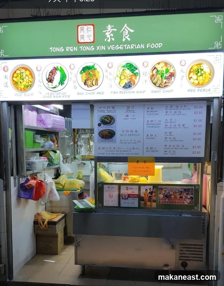 A brightly lit vegetarian food stall with a green awning displaying the business name and various food images. Below the images are menus with daily specials and prices. The stall has a display case with food items and promotional posters.
