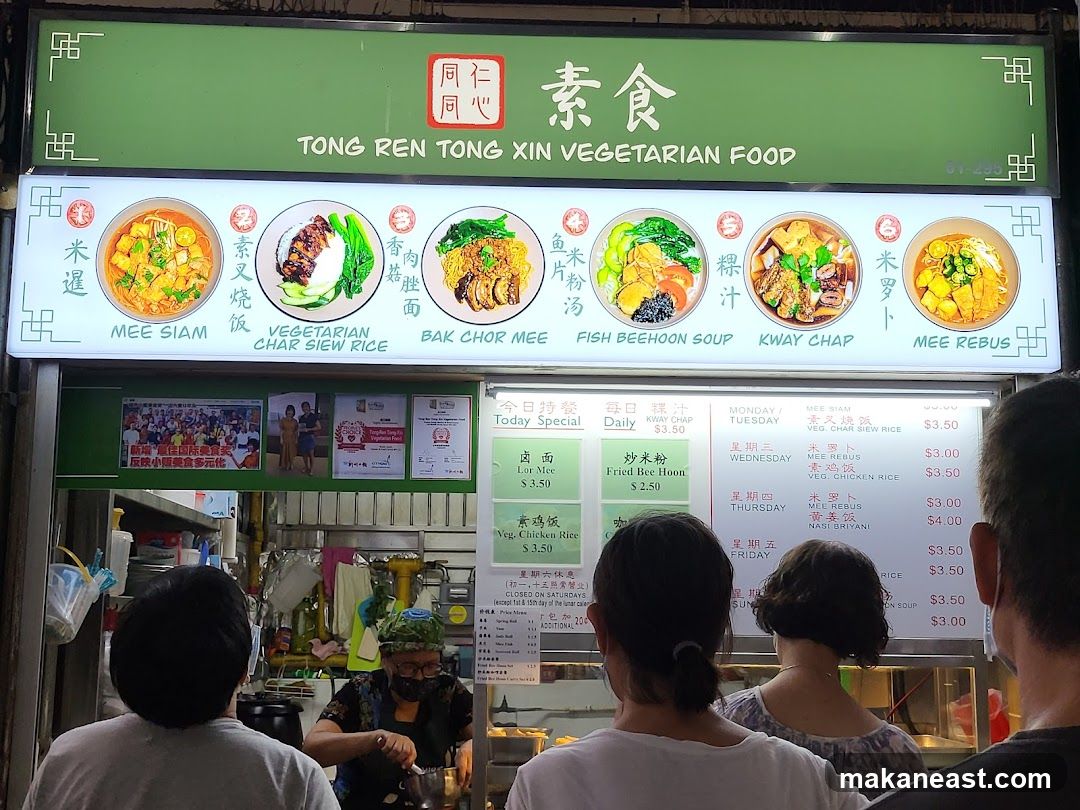 A brightly lit stall sign displays various vegetarian dishes with their names and images. Below the images, a menu board lists daily specials and prices for different days of the week. People are seen queuing at the stall.