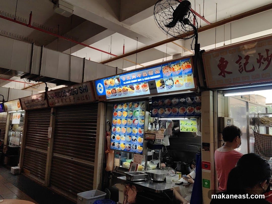 A dessert stall with various colorful images of desserts displayed on illuminated boards. The stall has a sign that reads "BLANCO COURT HOT & COLD DESSERT". Several shuttered stalls are visible to the left.