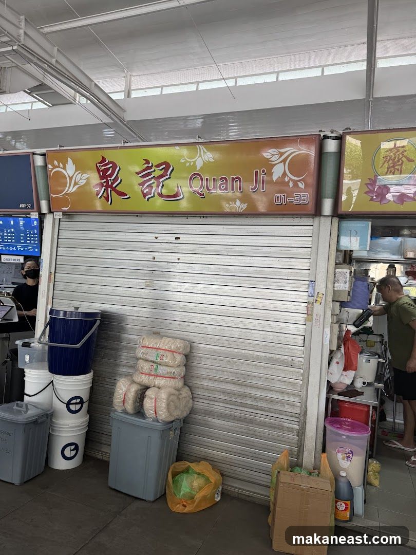 A closed shop front with a metal roller shutter. Above the shutter is a sign with the name "Quan Ji" in English and Chinese characters, along with the unit number "01-33". To the left of the sign, there is a smaller sign with text and numbers. To the right of the main sign, there is another sign with Chinese characters and a lotus flower design. In front of the shutter, there are stacks of buckets and bundles of what appear to be noodles. A person is visible on the right side of the image, near a counter with kitchen appliances.