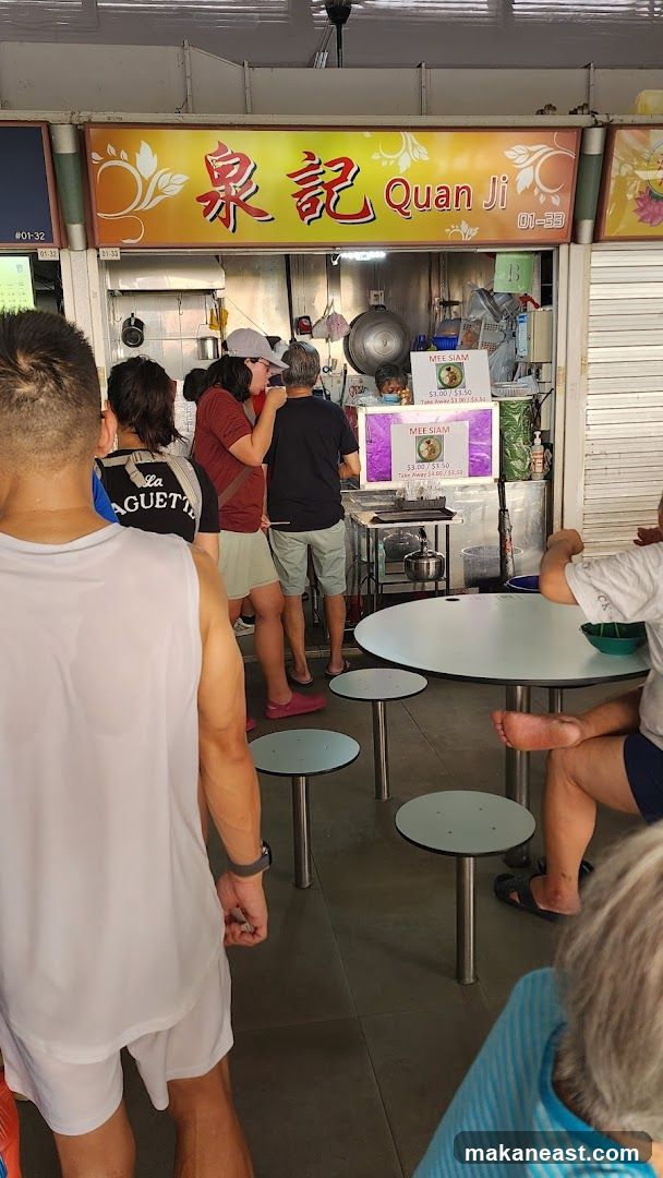 A busy food stall with a bright yellow sign that reads "泉記 Quan Ji" in Chinese and English. The stall is serving customers, with a line forming. There are tables and chairs set up in front of the stall. A menu board is visible, listing "MEE SIAM" with prices for dine-in and take away.