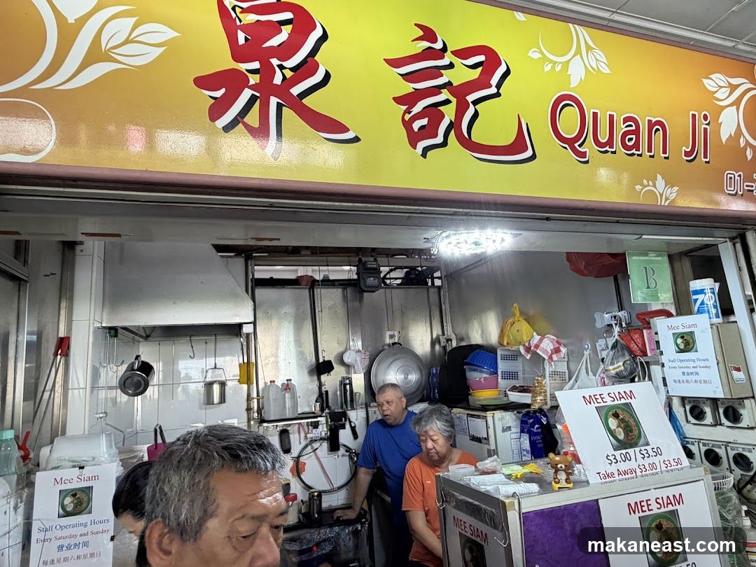 A food stall with a yellow awning displaying the name "Quan Ji" in Chinese characters and English. Below the awning is the stall's interior, showing kitchen equipment and staff. Signs for "Mee Siam" are visible, along with operating hours and prices.