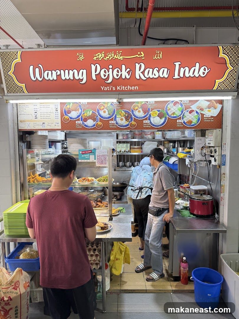 A food stall with a large red sign that reads "Warung Pojok Rasa Indo" and "Yati's Kitchen". Below the sign is a menu board displaying various dishes with pictures and prices. Several people are visible inside and in front of the stall, with food being prepared and served.