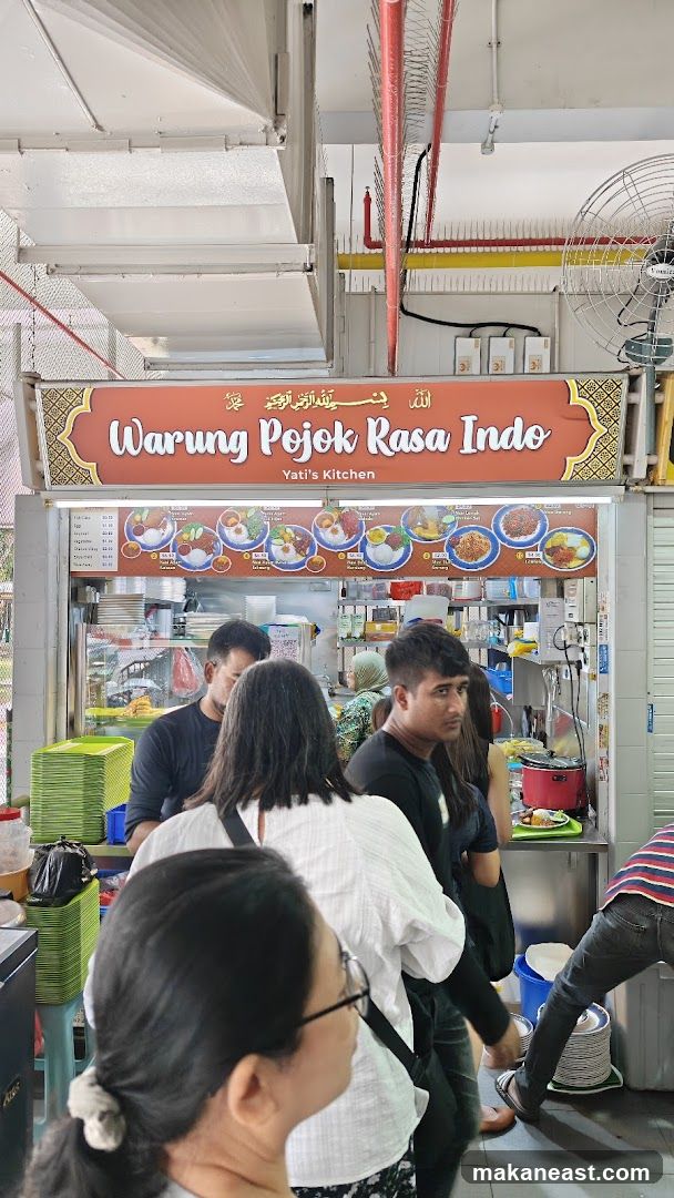 A food stall with a prominent sign displaying "Warung Pojok Rasa Indo" and "Yati's Kitchen". Below the sign is a menu board with pictures of various dishes. Several people are queuing to order food. The stall appears to be in an outdoor or semi-outdoor setting, possibly a hawker center.