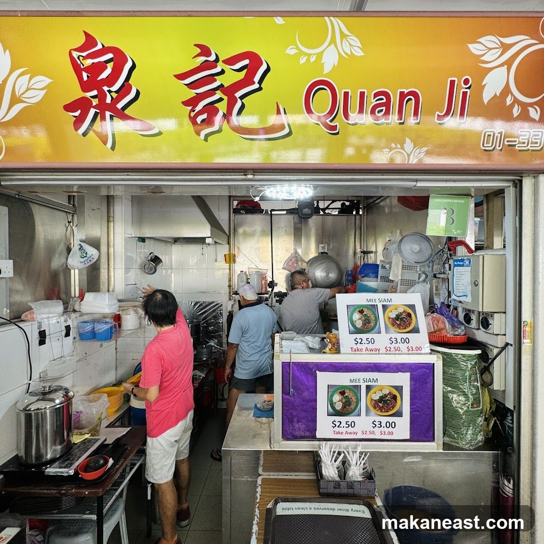 A food stall with a yellow sign featuring Chinese characters and the name "Quan Ji". The stall appears to be preparing food, with several people working inside. There are menu boards displaying "Mee Siam" with prices for dine-in and take away.