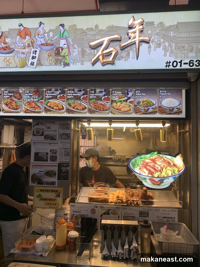 A food stall with a large illuminated sign featuring Chinese characters and illustrations of people preparing food. Below the sign is a menu display with images and prices of various dishes. Two staff members are visible behind the counter, one serving a customer. The stall has a selection of food items displayed, including fried items and bowls of rice.