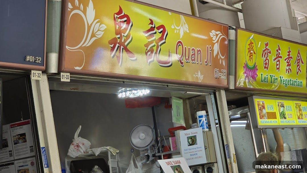 A close-up shot of two adjacent food stalls. The stall on the left has a sign with Chinese characters and "Quan Ji" in English. The stall on the right has Chinese characters and "Lei Yin Vegetarian" in English, with a lotus flower graphic. Below the signs, there are menu boards displaying food items and prices. The stall on the left has a sign for "Mee Siam" with operating hours.