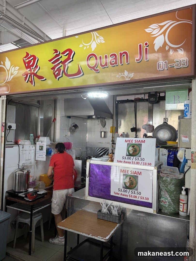 A food stall with a yellow and orange sign that reads "泉記 Quan Ji" and the stall number "01-33". A person in a red shirt and white shorts is working behind the counter. There are two signs advertising "MEE SIAM" for $3.00/$3.50, with a take away option for $3.00/$3.50.
