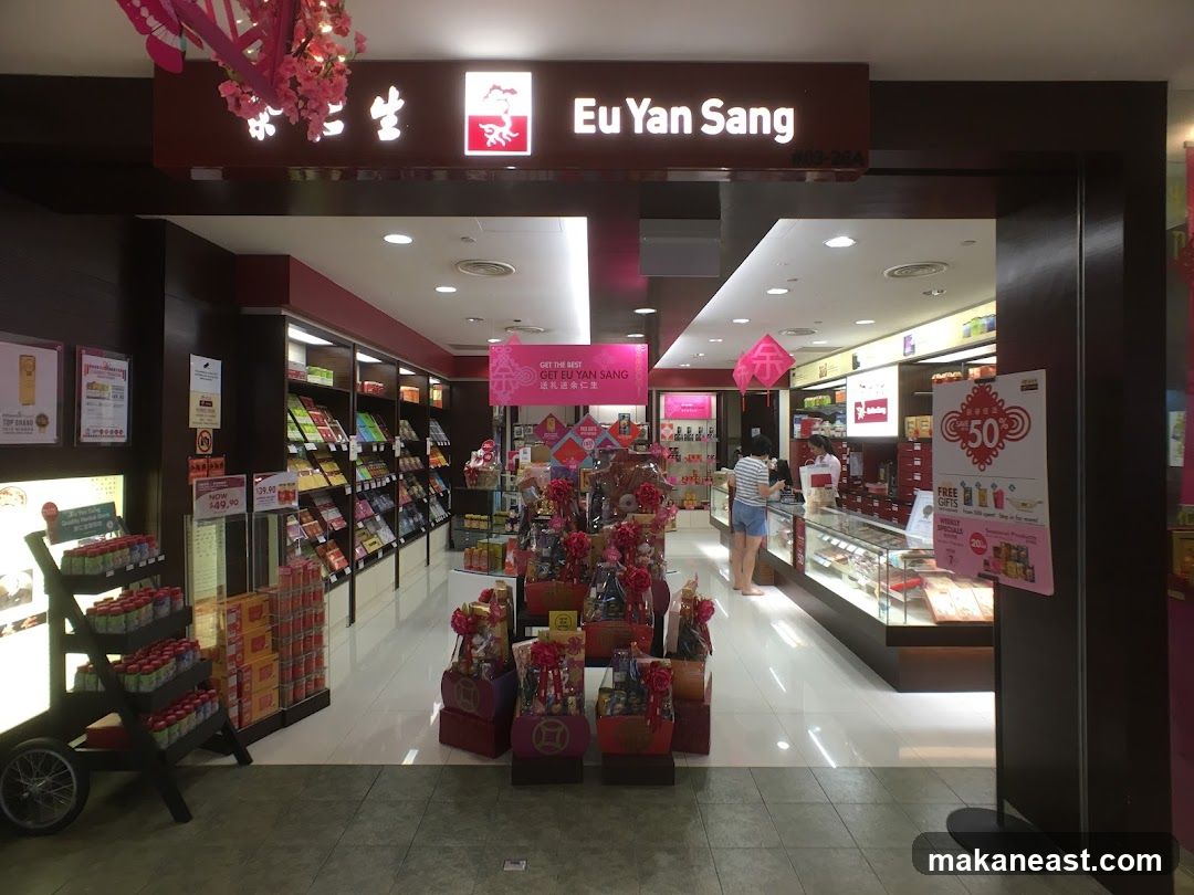 The exterior of a Eu Yan Sang store, with its prominent signage visible above the entrance. The store is well-stocked with various products displayed on shelves and in gift baskets. A promotional poster advertising 50% off and free gifts is placed near the entrance.