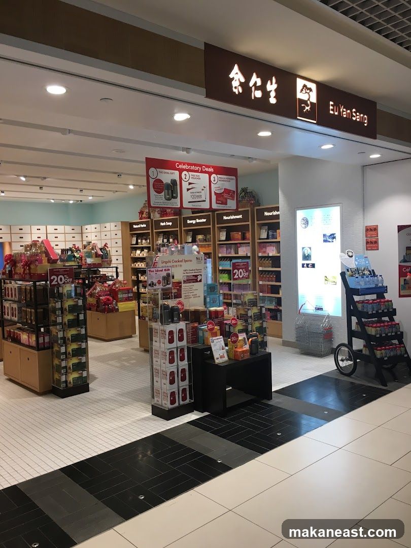The image shows the interior of a Eu Yan Sang store, a health and wellness shop. The store features shelves stocked with various products, including health supplements, traditional Chinese medicine, and gift sets. A prominent sign above the entrance reads "Eu Yan Sang" in English and Chinese characters. Promotional displays and signage are visible throughout the store, highlighting deals and product categories such as "Celebratory Deals," "Natural Herbs," "Energy Booster," and "Nourishment." The store has a clean and organized layout with white tiled floors and light-colored shelving units.