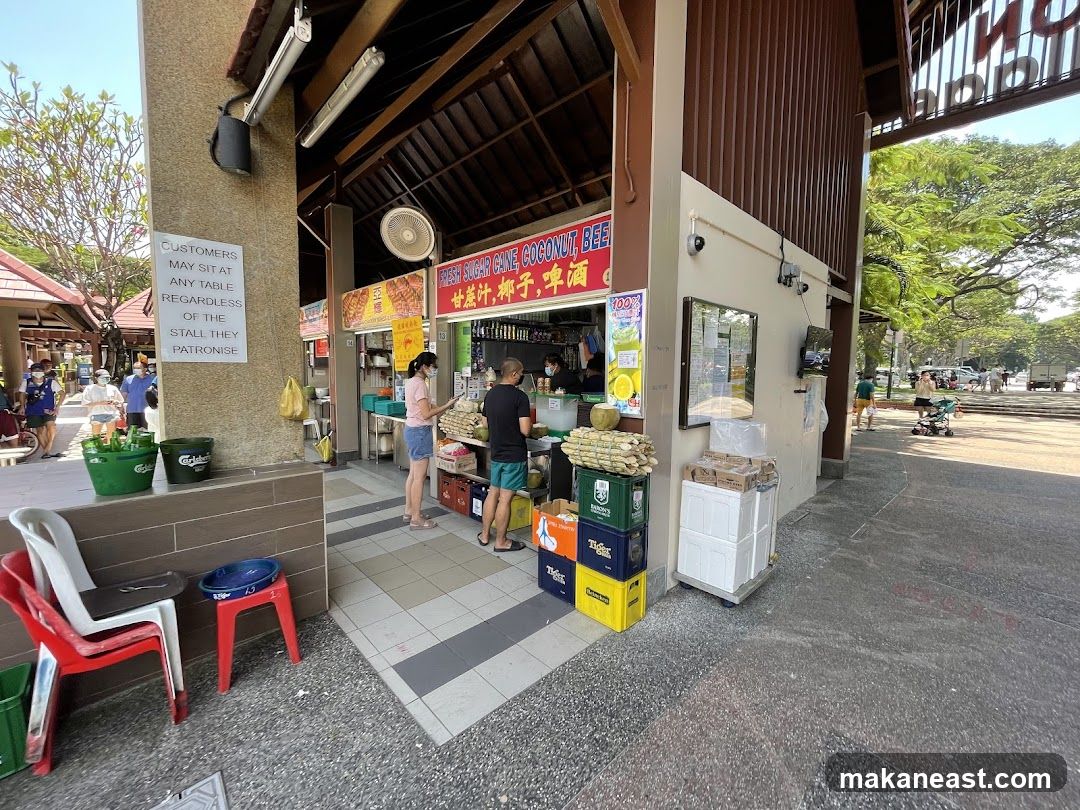 A street food stall selling fresh sugar cane juice, coconut, and beer. The stall has a sign with its offerings in English and Chinese. There are stacks of sugar cane, coconuts, and crates of drinks outside the stall. Customers are seen ordering and waiting for their drinks. A sign on a pillar states that customers may sit at any table regardless of the stall they patronise.