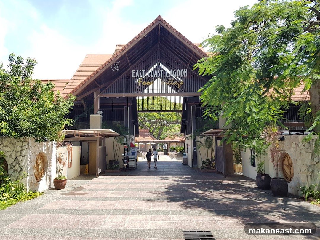 A wide shot of the entrance to East Coast Lagoon Food Village on a sunny day. The building has a large, open-air structure with a pitched, tiled roof supported by dark wooden beams. A prominent sign above the entrance reads "EAST COAST LAGOON VILLAGE" in illuminated letters. The facade is constructed with stone walls and features lush greenery on either side. A paved walkway leads towards the entrance, with several people walking in the distance.