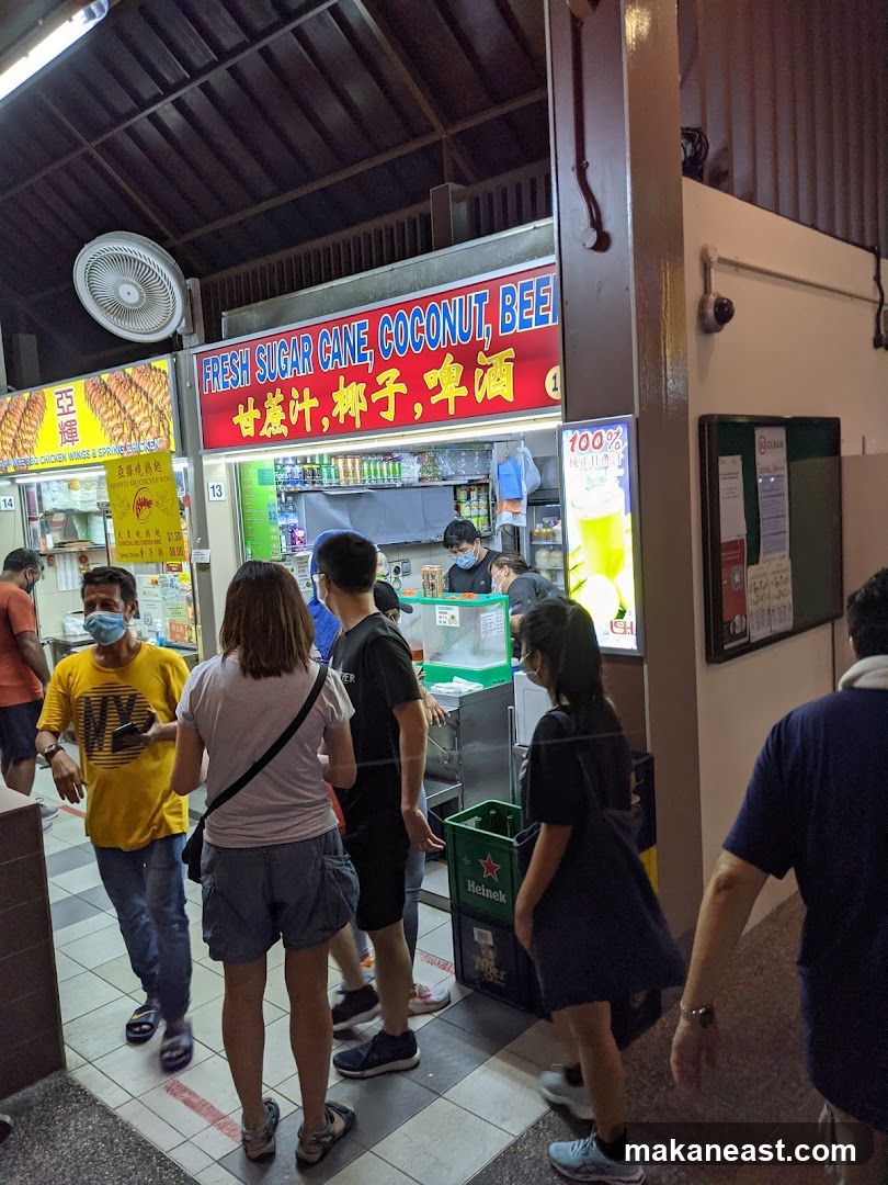 A stall with a red sign advertising "FRESH SUGAR CANE, COCONUT, BEER" in English and Chinese. People are queuing to order drinks. There are also signs for "CHICKEN WINGS & SPRING ROLLS".