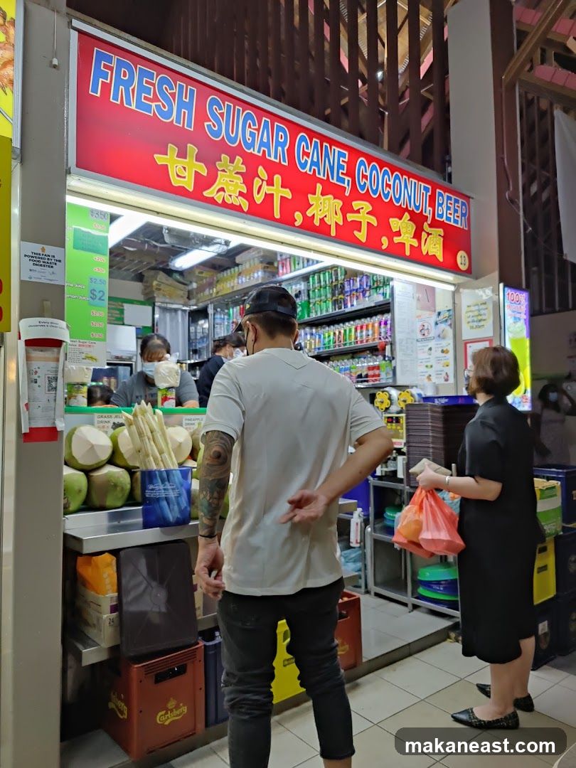A stall with a red sign displaying "FRESH SUGAR CANE, COCONUT, BEER" in English and Chinese. Coconuts and sugar cane stalks are displayed at the front. Several people are visible, including staff and customers. Shelves behind the counter are stocked with various drinks.