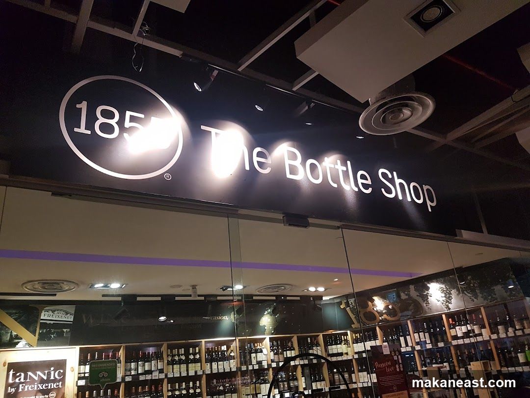 The exterior of a wine shop with a large sign above the entrance that reads "1855 The Bottle Shop". Through the glass doors, shelves stocked with wine bottles are visible.