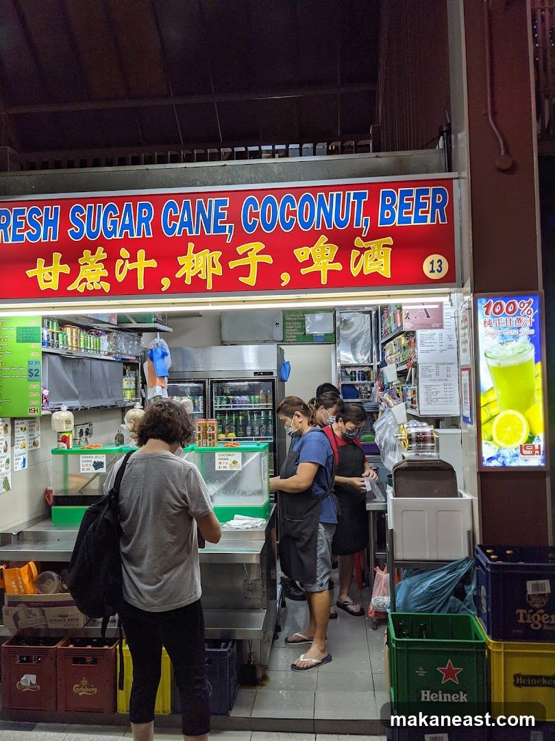 A stall with a large red sign displaying "FRESH SUGAR CANE, COCONUT, BEER" in English and Chinese. Several people are working behind the counter, preparing drinks. There are refrigerators stocked with beverages and containers of ingredients. Crates of beer bottles are visible in front of the stall. A promotional poster for sugarcane juice is on the right.