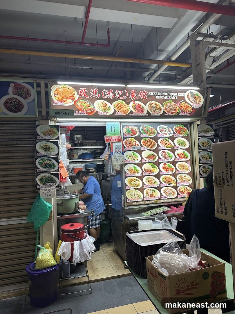 A food stall with a large illuminated sign displaying various dishes and the restaurant name. A man in a blue shirt is preparing food behind the counter. Stacks of trays and a box of produce are visible in the foreground.