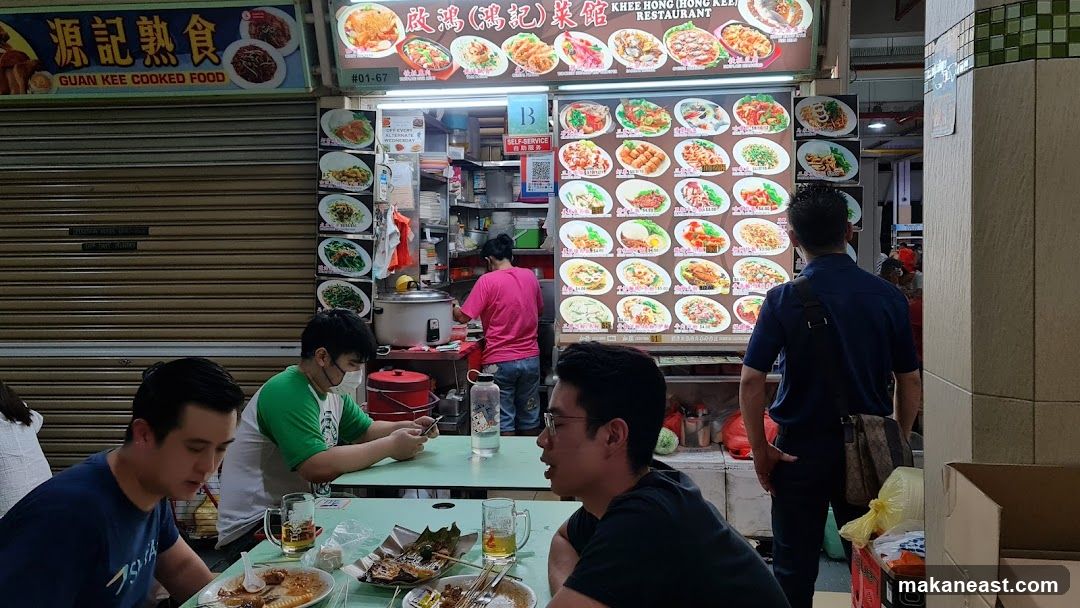 A food stall with a large menu board displaying various dishes. People are seated at tables in front of the stall, eating and looking at their phones. A person in a pink shirt is working behind the counter. The stall has signage in Chinese and English.