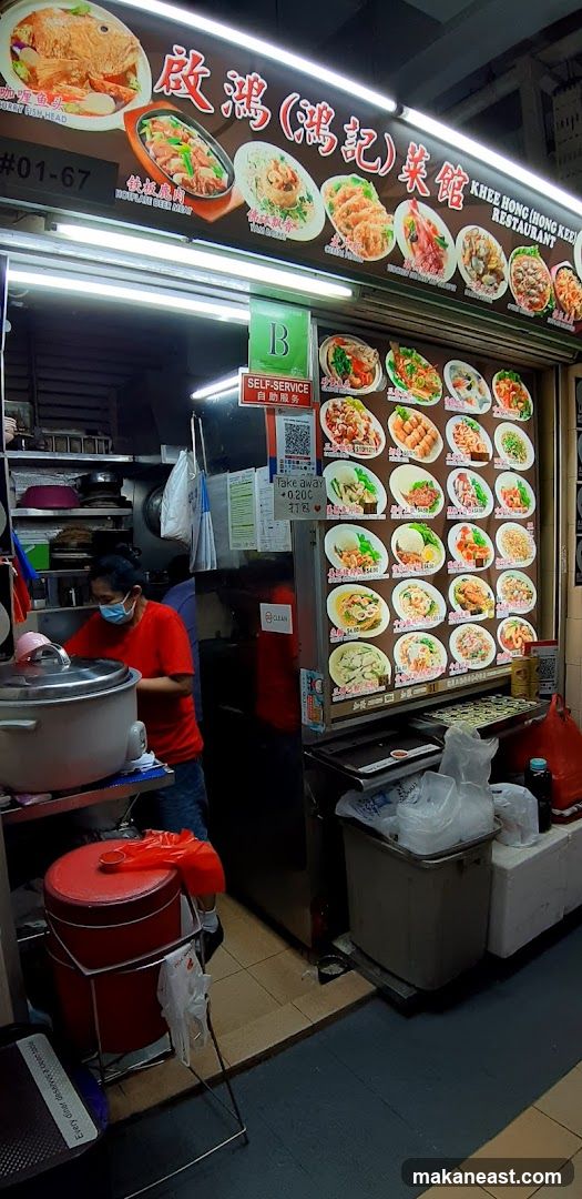 A food stall with a large illuminated sign displaying various dishes and the restaurant name. Below the sign, there are rows of pictures of food items with prices. A person wearing a red shirt and a mask is visible inside the stall, near a large rice cooker. There are also signs for self-service and takeaway.