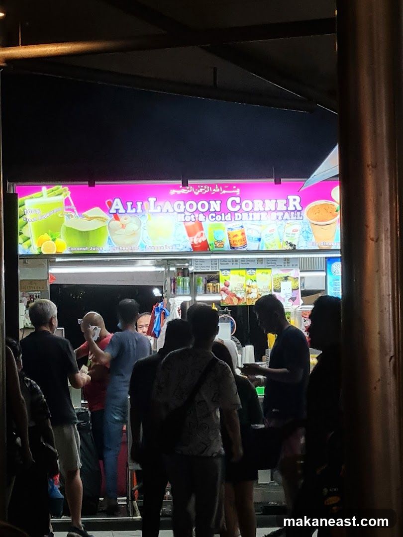 A brightly lit sign for "ALI LAGOON CORNER HOT & COLD DRINK STALL" is visible above a busy drink stall. The sign features colorful illustrations of various drinks, including coconut juice and what appears to be lychee juice. Several people are gathered at the stall, some ordering and others waiting for their drinks. The overall scene suggests a lively evening at a popular beverage stand.