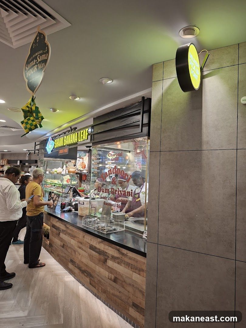 A view of the Shami Banana Leaf food stall with customers ordering. The stall features a sign with the name "SHAMI BANANA LEAF" and displays food items behind a glass counter. There are also signs indicating "Charcoal Grill" and "Dum Briyani with slow-cooked Basmati Rice". A circular sign on the wall reads "DELIVER PICK UP POINT".