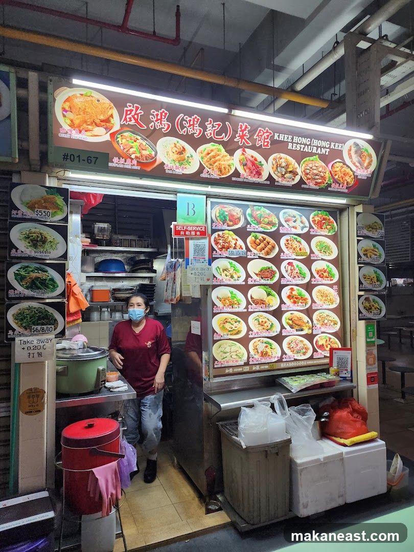 A food stall with a large illuminated sign displaying various dishes and prices. A woman wearing a mask stands behind the counter. The stall has a menu board with pictures of food items and prices.
