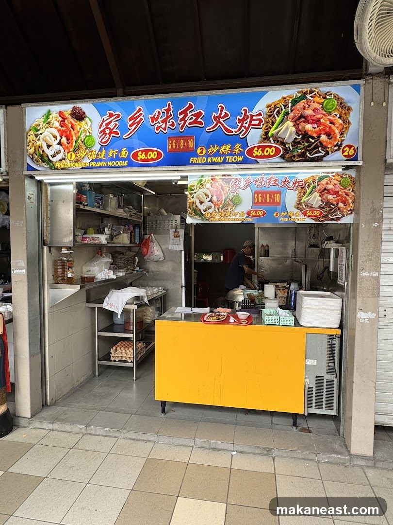 A food stall with a bright yellow counter and a large overhead sign displaying menu items. The sign features images of noodle dishes and their prices. The stall appears to be open with a person working inside.