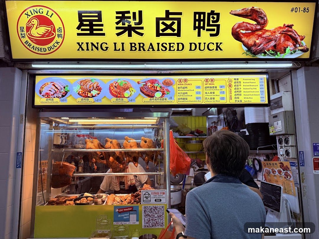 A bright yellow awning with the name "XING LI BRAISED DUCK" in English and Chinese characters. Below the awning, a display case shows several roasted ducks and other meats. A menu board with pictures and prices of various duck dishes is visible above the display case. A person is standing in front of the display, looking at their phone.