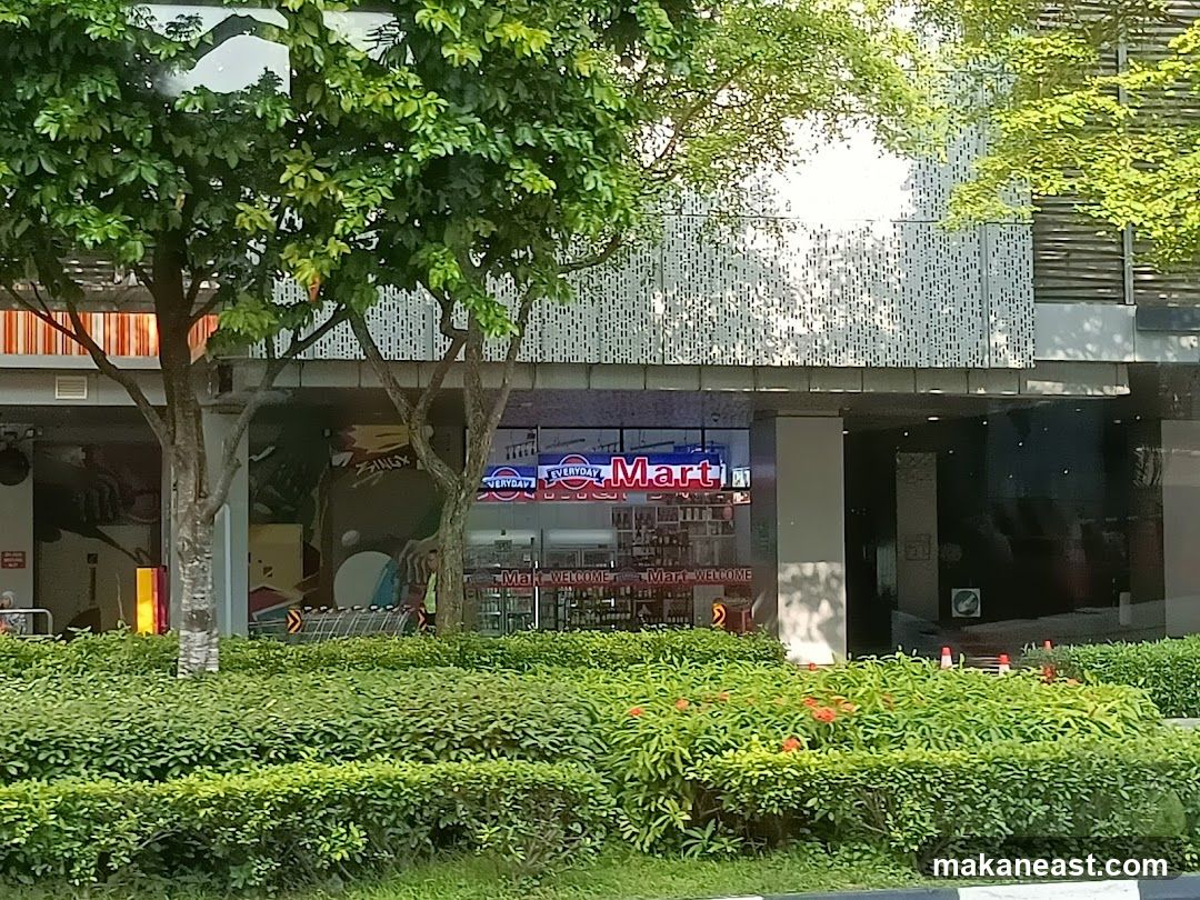 The storefront of a convenience store called Everyday Mart. The store has large glass windows displaying shelves stocked with various products. The store's name is prominently displayed in neon red and blue letters above the entrance. A green hedge and trees are in front of the store.