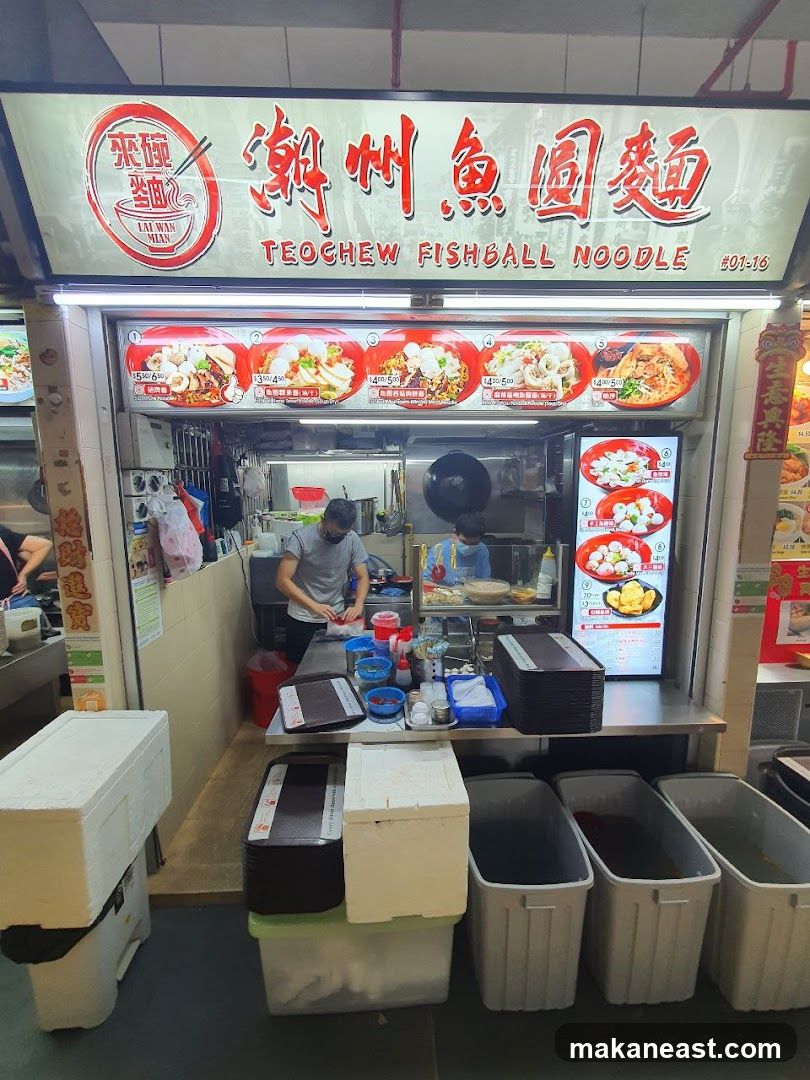 A food stall with a prominent sign displaying "Teochew Fishball Noodle" in English and Chinese. Below the sign are several images of noodle dishes with prices. Two staff members are visible inside the stall, one preparing food and the other serving. In front of the stall are several large containers and stacked boxes.