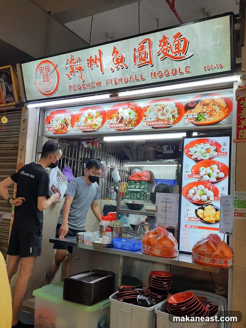 A street food stall with a large illuminated sign displaying "Teochew Fishball Noodle" in English and Chinese. Below the sign are images of various noodle dishes. Two men are visible at the counter, one serving and one ordering. Stacks of bowls and containers are visible in the foreground.