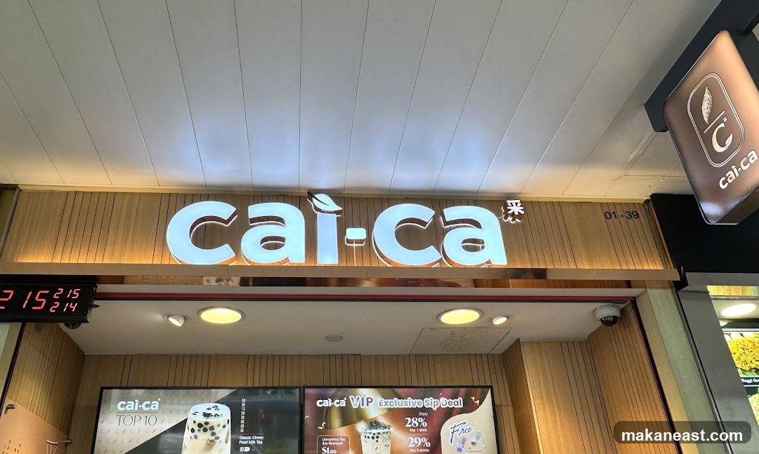 The image shows the exterior signage of a Cai-ca store. A large illuminated sign with the Cai-ca logo and name is prominently displayed. Below the main sign, there are posters advertising promotions and menu items, including a "TOP 10" list and a "VIP Exclusive Sip Deal." A digital clock showing "215 214" is visible on the left. A smaller hanging sign with the Cai-ca logo is also present.
