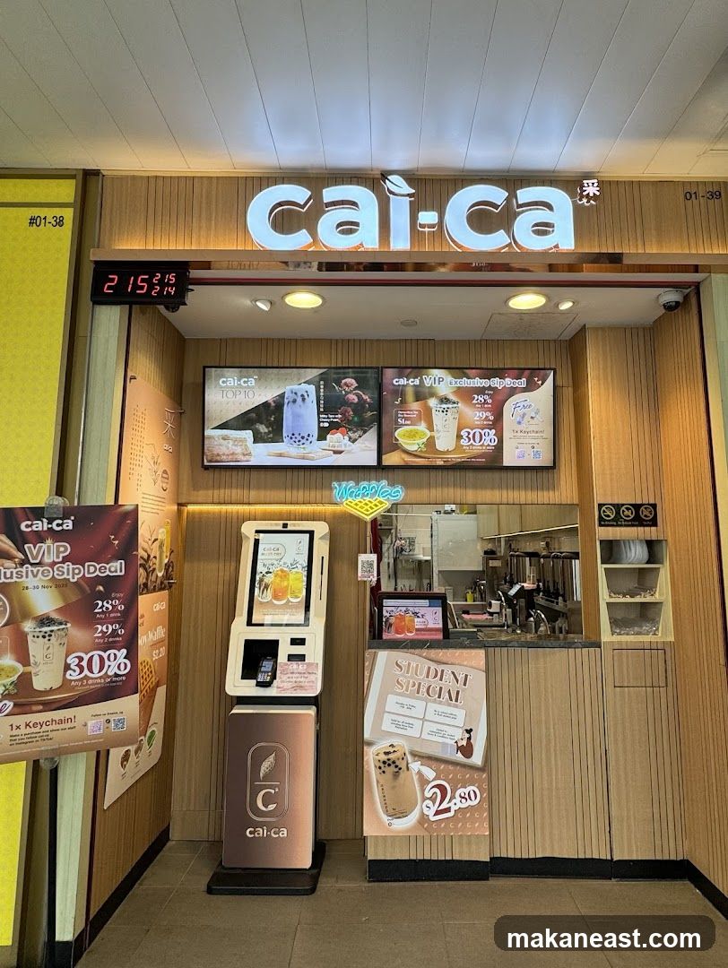 The storefront of a Cai-Ca bubble tea shop. A large illuminated sign with the shop's name is visible above the entrance. To the left, a digital clock displays the time. There are promotional posters and a self-ordering kiosk. The counter area with beverage preparation equipment is visible behind the counter.
