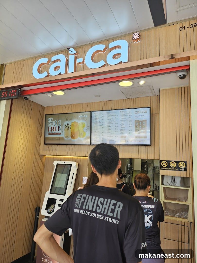 The exterior of a Cai-Ca bubble tea shop with its prominent signage. A digital menu board displays promotions and menu items. Customers are seen queuing, with one person in the foreground wearing a black t-shirt with "FINISHER ARMY READY SOLDIER STRONG" printed on the back. A self-ordering kiosk is also visible.