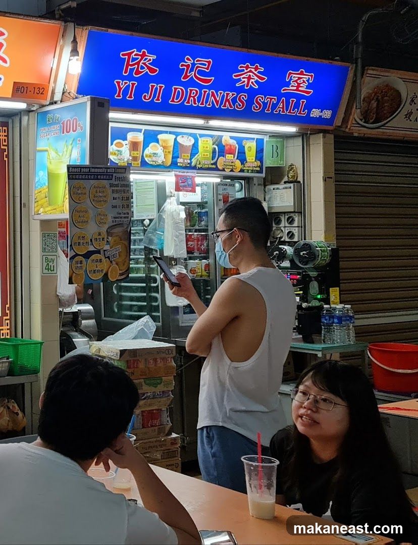 A street-level view of a drinks stall with a blue sign that reads "YI JI DRINKS STALL" in English and Chinese. Below the main sign is a menu board displaying various drinks with prices. A man wearing glasses and a face mask is standing in front of the stall, looking at his phone. Two customers are seated at a table in the foreground. Stacks of boxes are visible to the left of the stall.