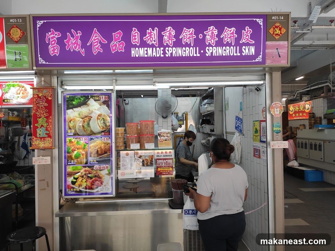 A food stall with a purple awning displaying Chinese characters and the text "HOMEMADE SPRINGROLL - SPRINGROLL SKIN". There are pictures of food items on display, including spring rolls, fried items, and a salad. Two staff members are visible behind the counter. The stall appears to be in an indoor market or food court.