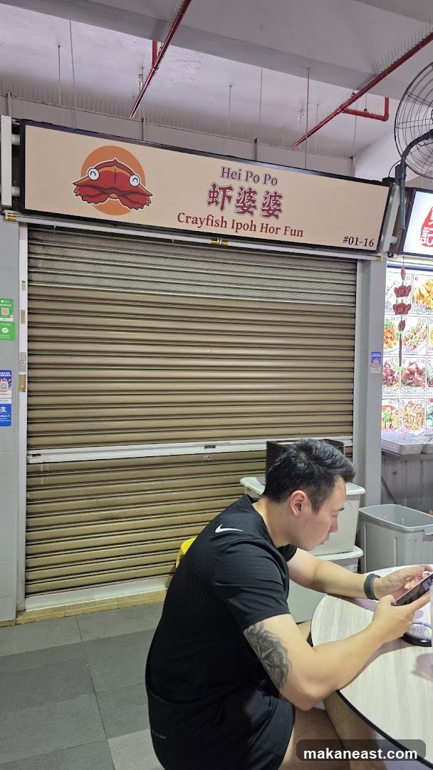 The exterior of a food stall with a closed metal shutter. Above the shutter is a sign with a cartoon crab and the text "Hei Po Po 虾婆婆 Crayfish Ipoh Hor Fun #01-16". To the right of the stall, a menu board with pictures of food is visible. A man is sitting at a table in front of the stall, looking at his phone.