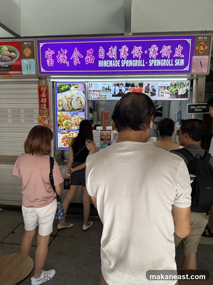 A food stall with a purple sign that reads "富城食品 自制醇解·春餅皮 HOMEMADE SPRINGROLL · SPRINGROLL SKIN". Below the sign is a menu display with pictures of spring rolls and other food items. Several people are queuing in front of the stall.