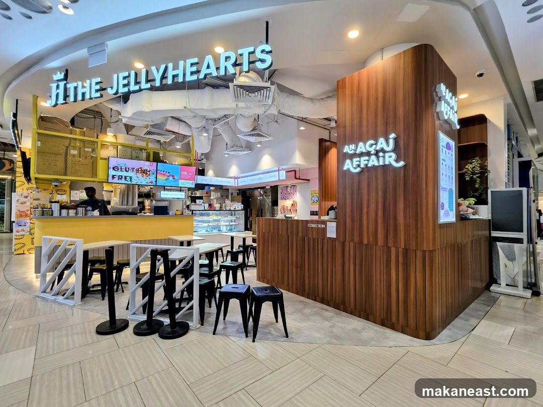 A brightly lit food stall with two distinct brands. On the left, "THE JELLYHEARTS" is displayed in large, illuminated letters above a yellow counter. A screen shows "GLUTEN FREE". On the right, "AÇAÍ AFFAIR" is also illuminated on a wooden facade. Seating areas with tables and stools are visible in front of the stalls.