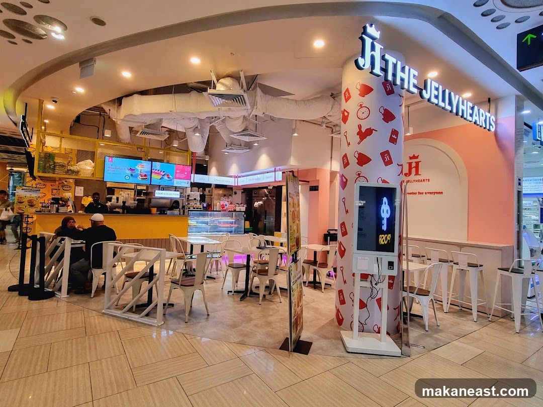 A brightly lit dessert shop with a yellow counter and pink accents. The shop's name, "THE JELLYHEARTS," is prominently displayed in large white letters above the entrance. A digital menu board is visible behind the counter, showing images of cakes and desserts. Several tables and chairs are set up for customers. A decorative pillar features the shop's logo and a whimsical pattern of keys and rabbits.