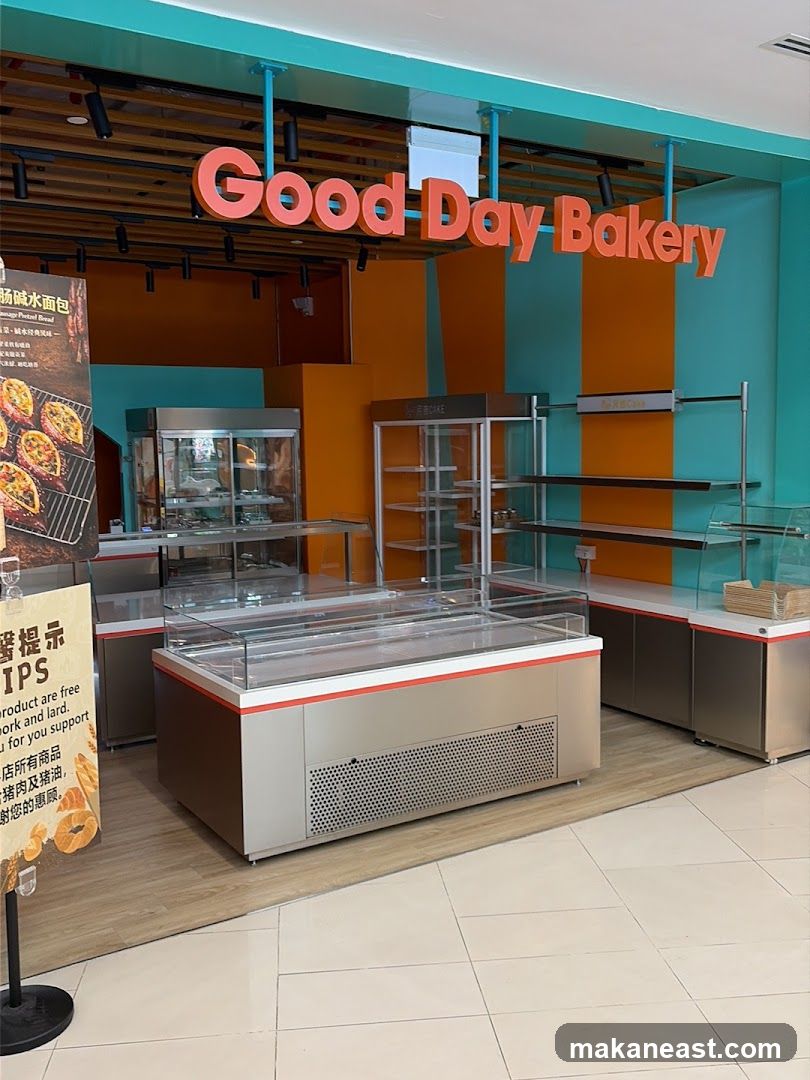 The exterior of a bakery with a large orange sign that reads "Good Day Bakery". There are display cases and shelves visible inside the shop. A poster with Chinese text and images of bread is to the left of the entrance.